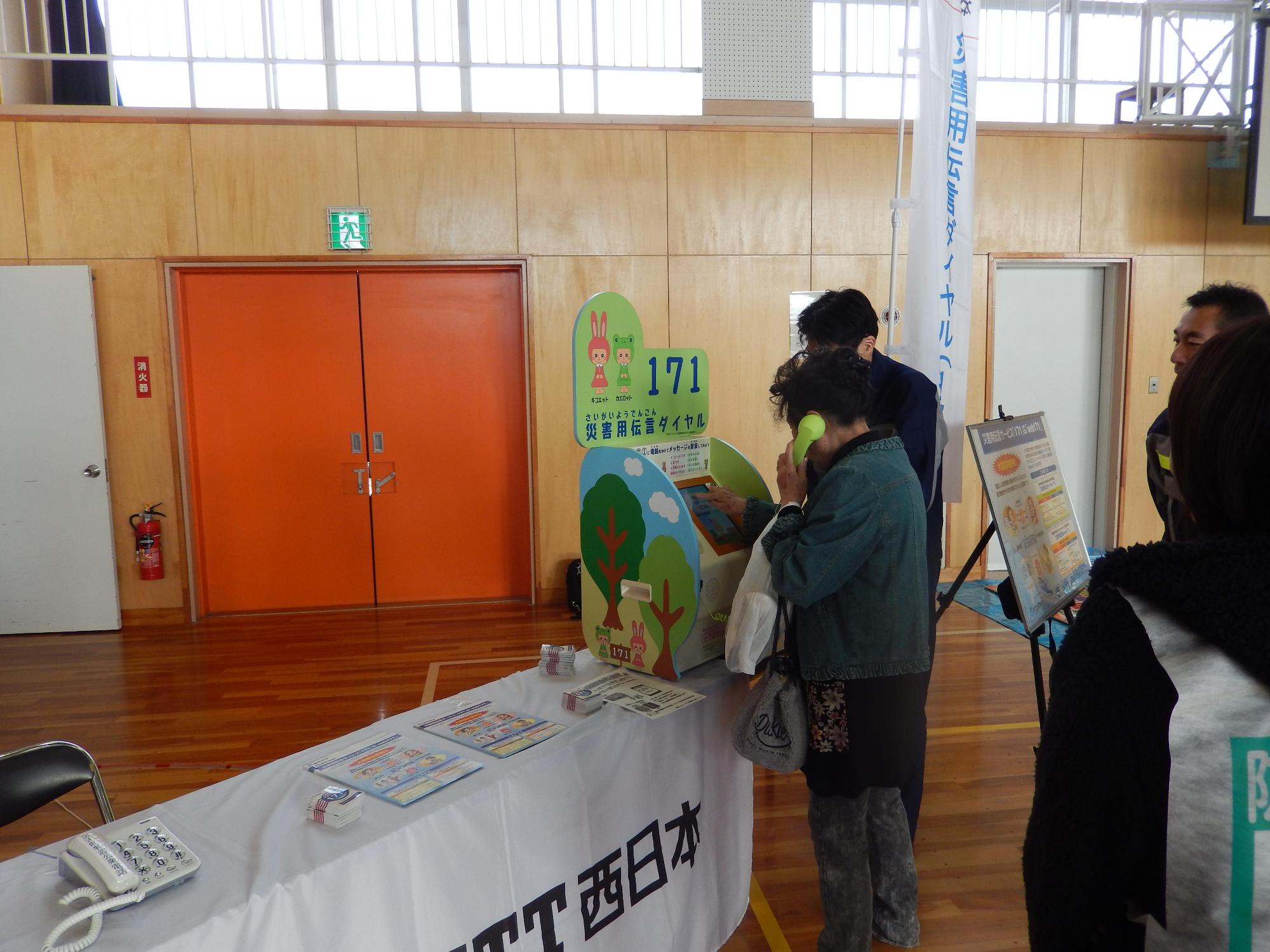 体育館の長机に固定電話と資料が並べられている横に設置されたNTT西日本の電話で電話をしている参加者を見ているスタッフや他の参加者たちの写真