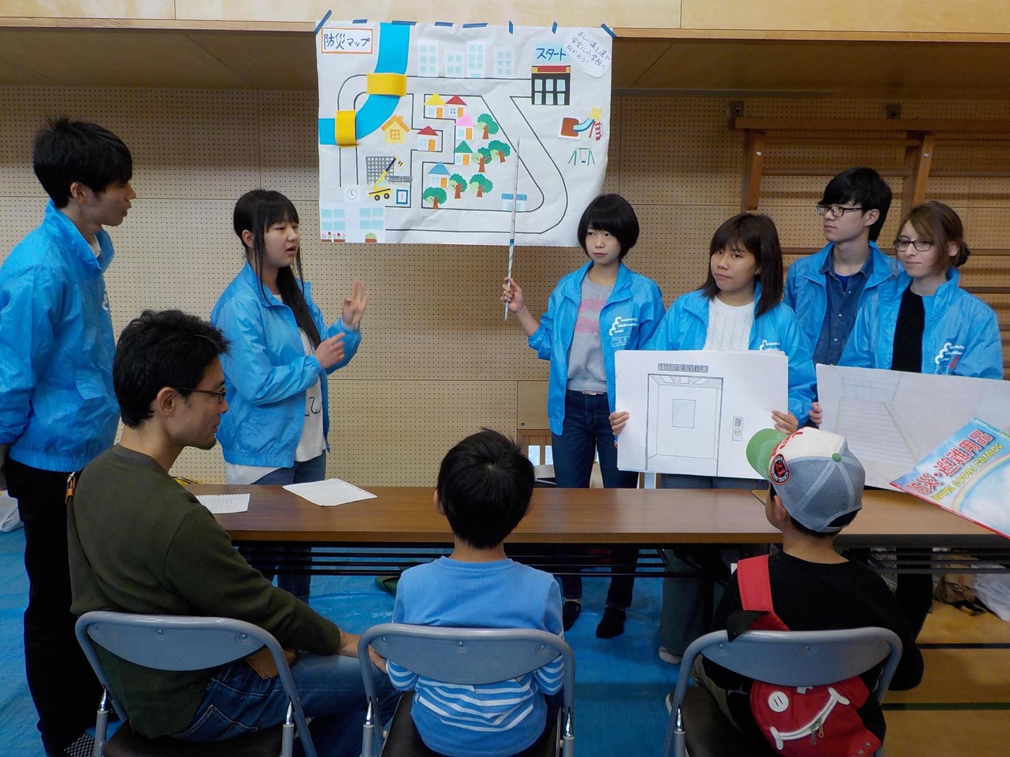 壁に掲示された防災マップの方を1人の女子大学生が指揮棒で指し、他の女子大学生が説明するのを椅子に座って聞いている親子たちの写真