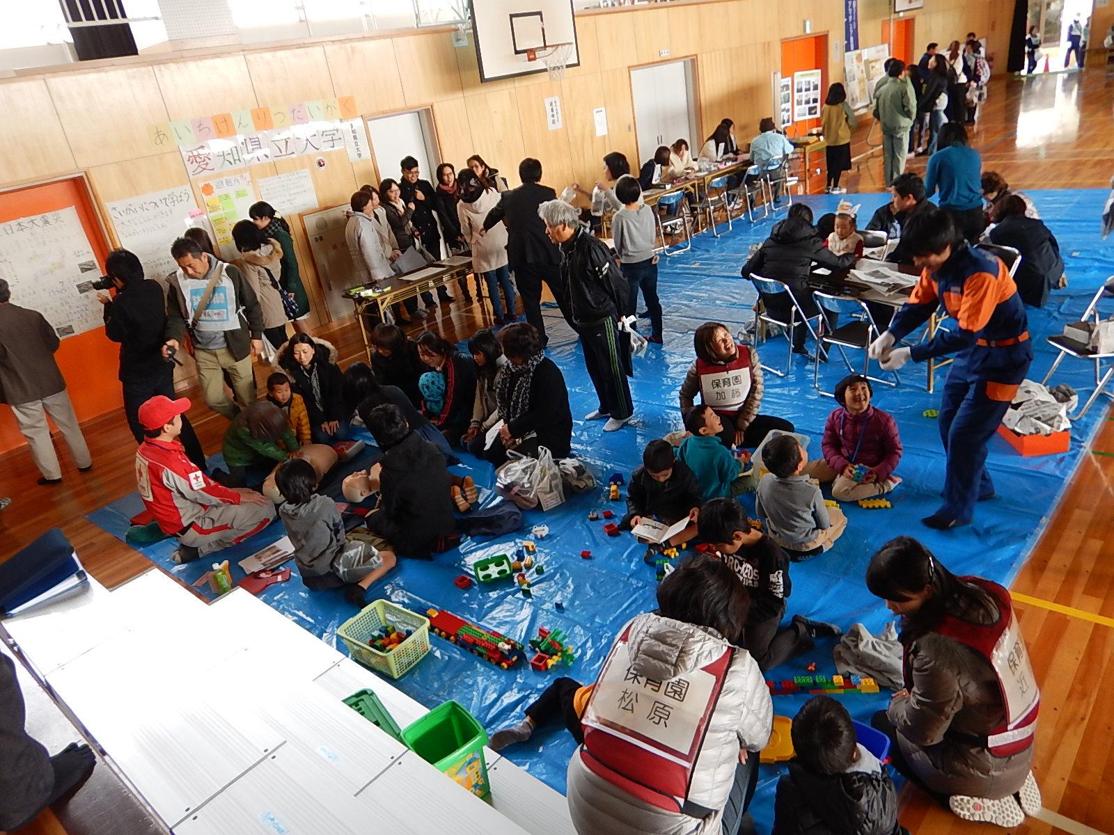 体育館に敷かれたブルーシートの上でおもちゃを広げ小さい子供と一緒に訓練をしている様子の写真