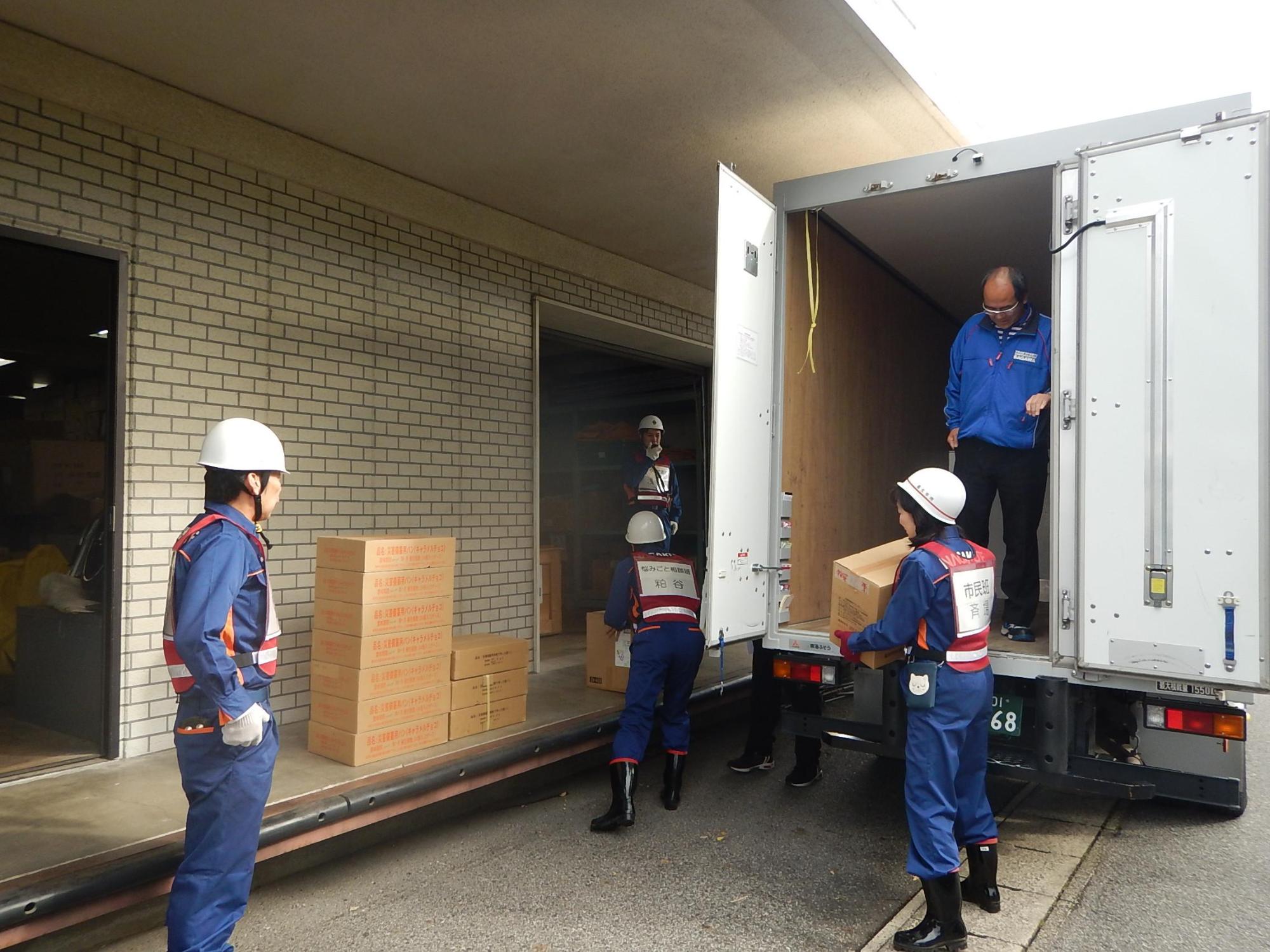 青い作業服に白いヘルメットを被っている市職員たちがトラックから物資を荷下ろしする様子を青いジャンバーを着た佐川急便株式会社のスタッフがトラックの荷台から見ている写真