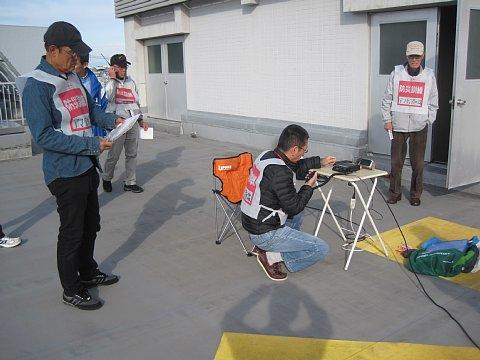 (市役所)屋外でアマチュア無線クラブによる情報収集・伝達訓練でテーブルの上で無線機を操作しているクラブ員と、他のクラブ員がトランシーバーで無線を聞いている写真