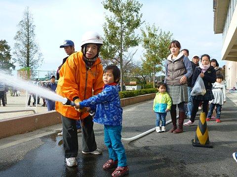 女性の担当者が支えながら男の子が放水訓練を行っており、その後ろで列をなして参加者が待っている写真
