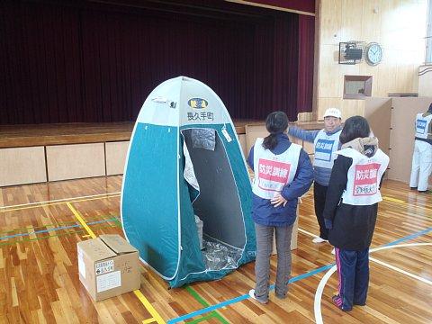 体育館の中で、薄水色の簡易トイレの展示の説明を参加者の2名の女性が聞いている写真