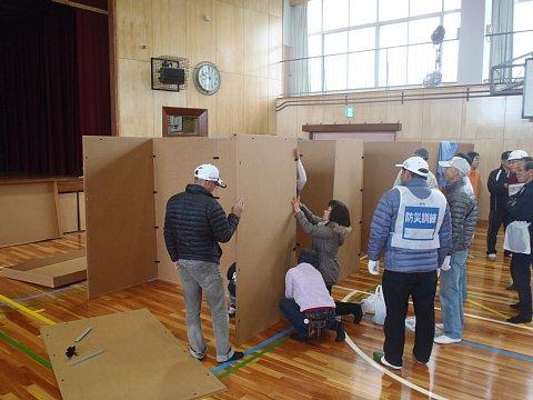体育館の中で、スタッフに教わりながら参加者の女性が段ボールを間仕切りのセッティングの訓練を行っている写真