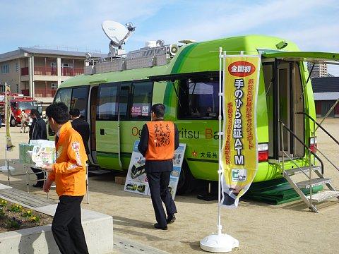 校庭の隅に、パラボナアンテナが付いていて車体が薄緑色の災害時用ATM搭載車を展示している写真
