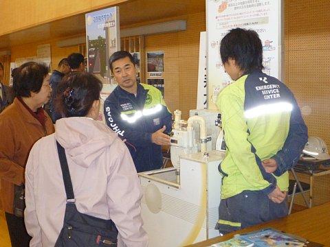 紺色の作業着を着ている男性のガス会社の担当者2名が、避難訓練に参加した女性2名に熱心に説明している写真