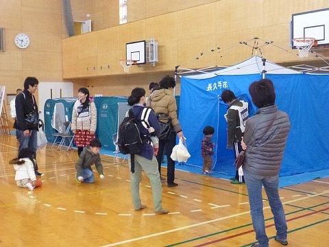体育館の中に長久手市と書かれた青色の大きな簡易テントの入り口を開けて見ている親子と、遠巻きに見ている参加者の写真