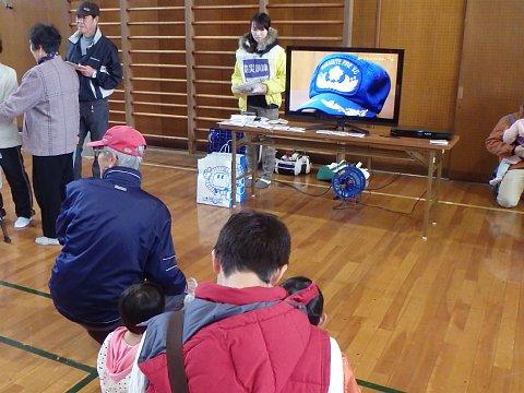 長机の上に災害時放送が映し出されている大きなテレビが設置しており、親子の参加者や高齢の男性が見ている写真