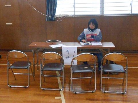体育館内に設置された長机に紙で「手話コーナー」と貼られた右側に女性が座り、机の前にはパイプ椅子が4脚並んでいる写真