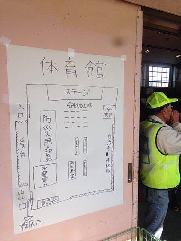(南小)会場配置図が体育館の入り口に掲示されている横に黄色い帽子に黄色いベストを着たスタッフが写っている写真