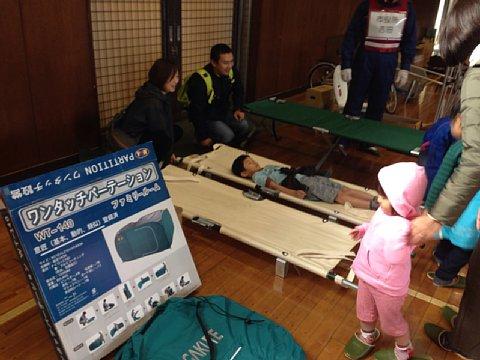 (南小)防災資機材の展示コーナーにある担架に子供が寝ているそばに両親がいる写真