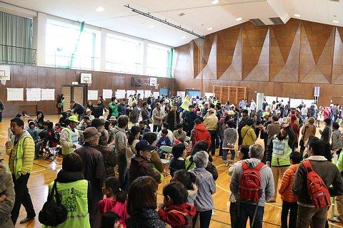 (南小)リュックを背負った参加者たちや黄色いベストを着たスタッフたちが体育館内に集まっている写真