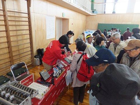 (長小)NTTドコモによる災害用伝言板体験訓練ブースの受付に携帯電話やスマートフォンが並べられており、赤いジャンバーを着たスタッフより説明を受ける参加者たちの写真