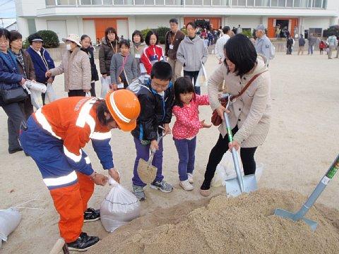 (長小)土のう袋を持っている長久手建設業協会のスタッフの横でスコップを使って砂を入れようとしている参加者親子を周りの参加者が見ている写真