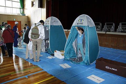 (長小)体育館の防災資機材の展示コーナーにて簡易トイレに入る子供や、レスキューセットが展示されている写真
