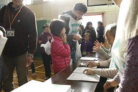 (長小)体育館の入口付近に設置された避難所受付で受付をする親子と並ぶ参加者たちの写真