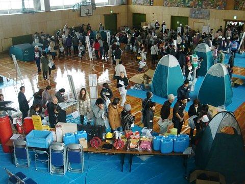 (西小)体育館内にてポリタンク、懐中電灯などの防災グッズが並べられているテーブルの前に参加者たちが資料を持って並んでおり、奥に簡易トイレなどの防災資機材展示コーナーに人々が集まっている写真