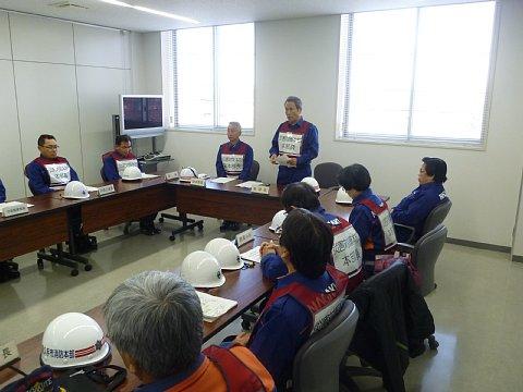 (市役所)テーブルの上に資料とヘルメットが置いてある会議室で市長の話をテーブルに座って聞いている市職員たちの写真