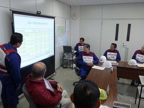 (市役所)テーブルの上に資料とヘルメットが置いてある会議室でスクリーンを前に市職員が説明をしており、他の職員たちが説明を聞いている写真