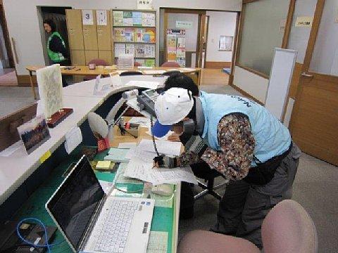 (福祉の家)カウンター受付の机にパソコンが置いてある横で電話応対しながらメモを取る水色のベストを着たボランティアスタッフの写真