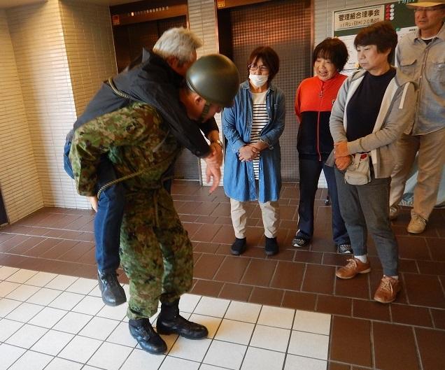 4名の参加者が見守る中迷彩服を着た自衛隊員が年配の男性を背負い救急搬送訓練をしている様子の写真
