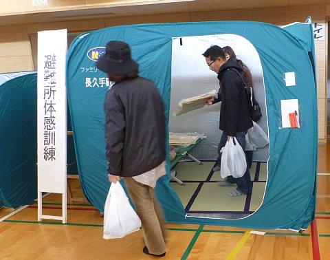 「避難所体感訓練」と書かれている看板の右側に、四角の形で右半分がチャックが空いて出入りが出来るテントの中に参加者が入っている写真