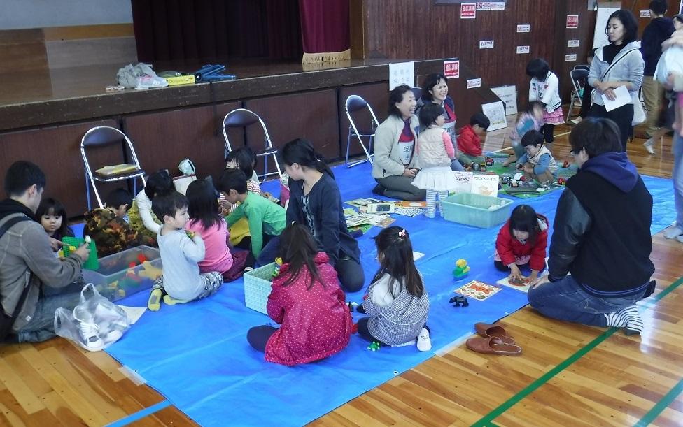体育館の一部にブルーシートを敷きおもちゃなどを広げて子供たちが遊んでいる避難所保育園の様子の写真