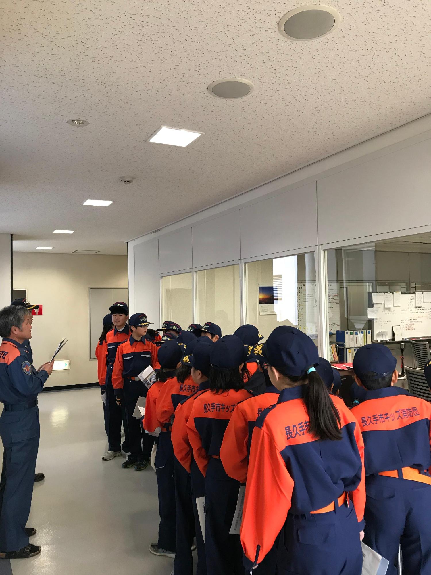 視察研修にて消防団員の説明を聞きながらガラス越しに指令センターを見学するキッズ消防団員たちの写真