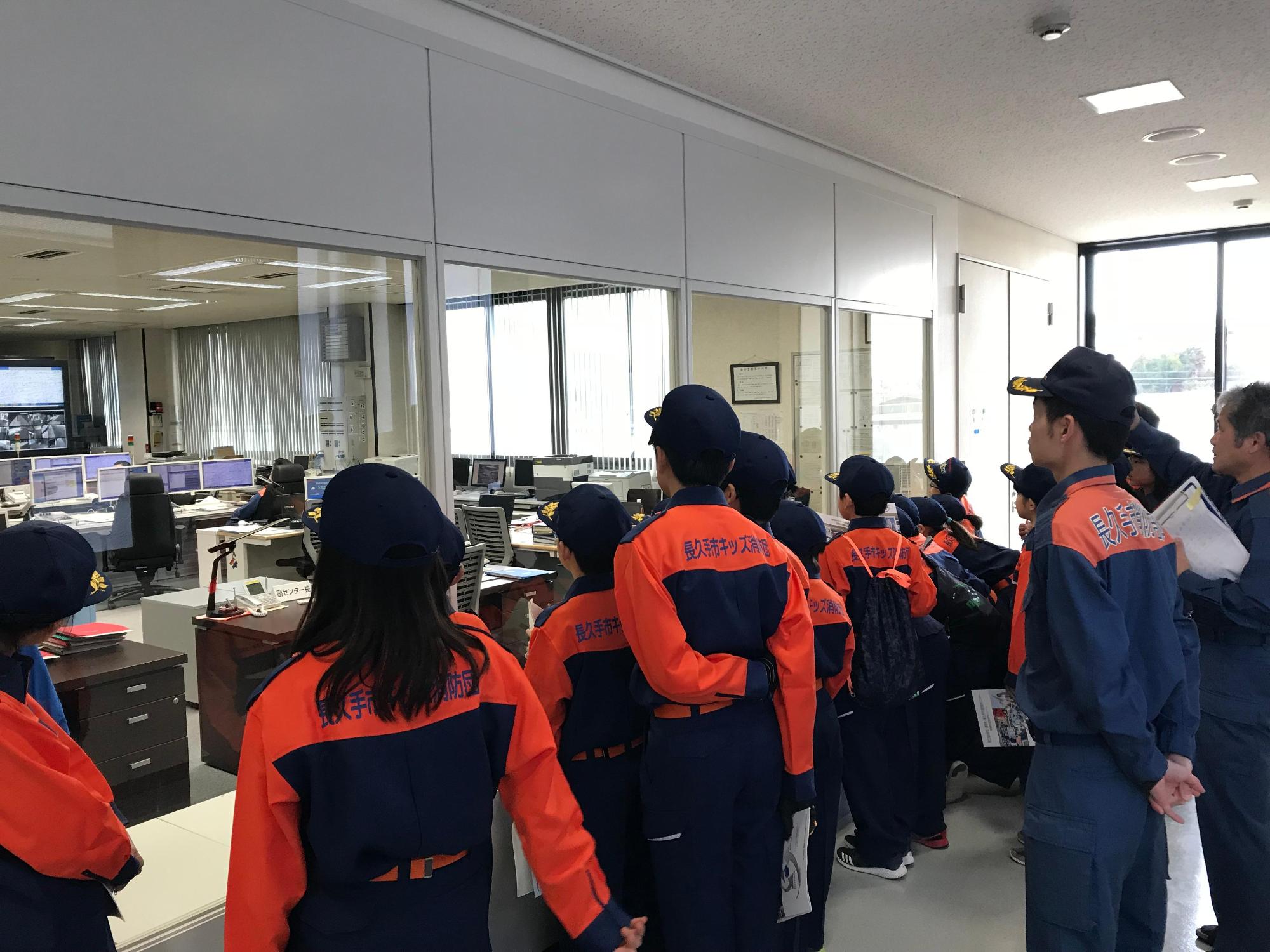 視察研修にてガラス越しに指令センターのモニターを見るキッズ消防団員たちの写真