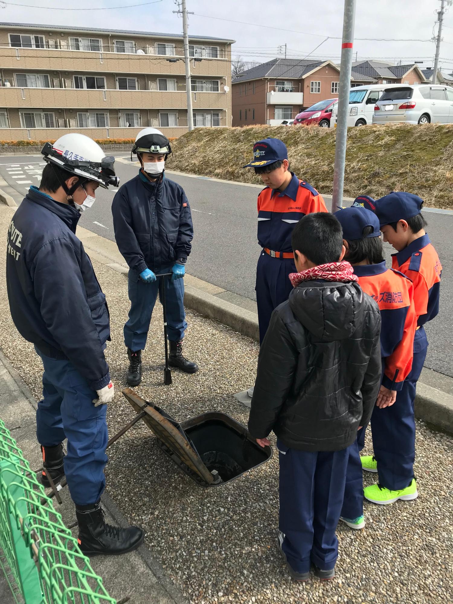 歩道に設定された消火栓の蓋を消防署員が開けて、キッズ消防団員たちに中を見せている写真
