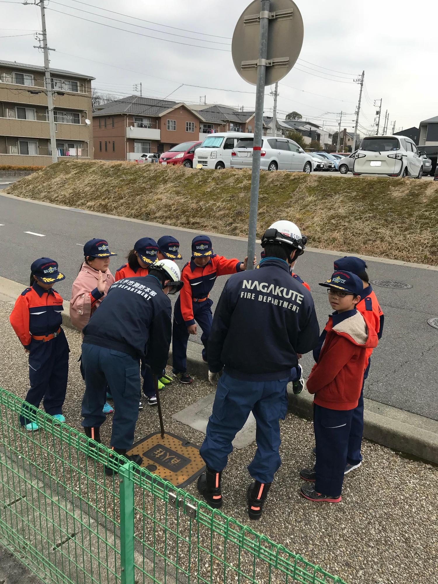歩道に設定された消火栓の蓋を消防署員が棒で締めている様子をキッズ消防団員たちが見ている写真