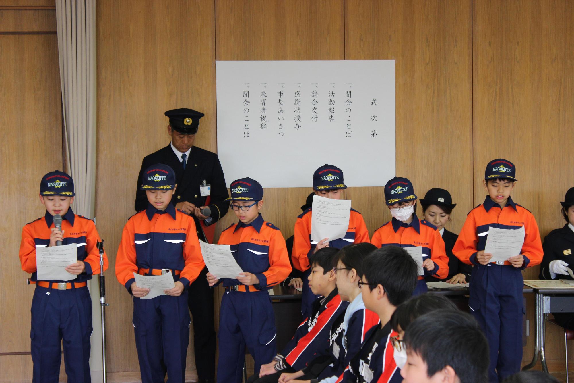 式次第の前で消防団長と、キッズ消防団員が整列して資料を読み上げている写真