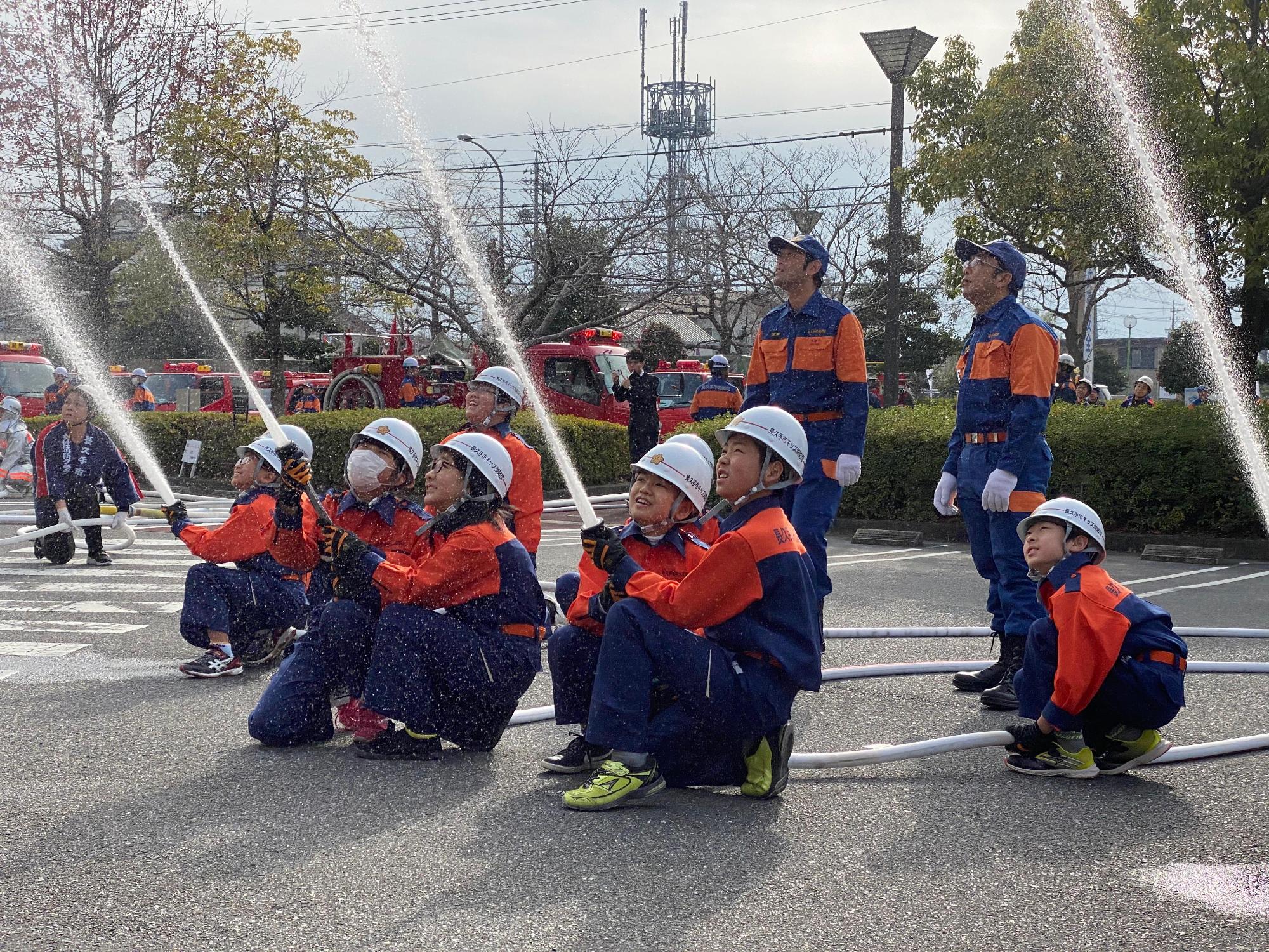 消防車両が止まっている駐車場で長久手市キッズ消防団員が2名1組になって一斉放水しているのを見守っている消防団員の写真