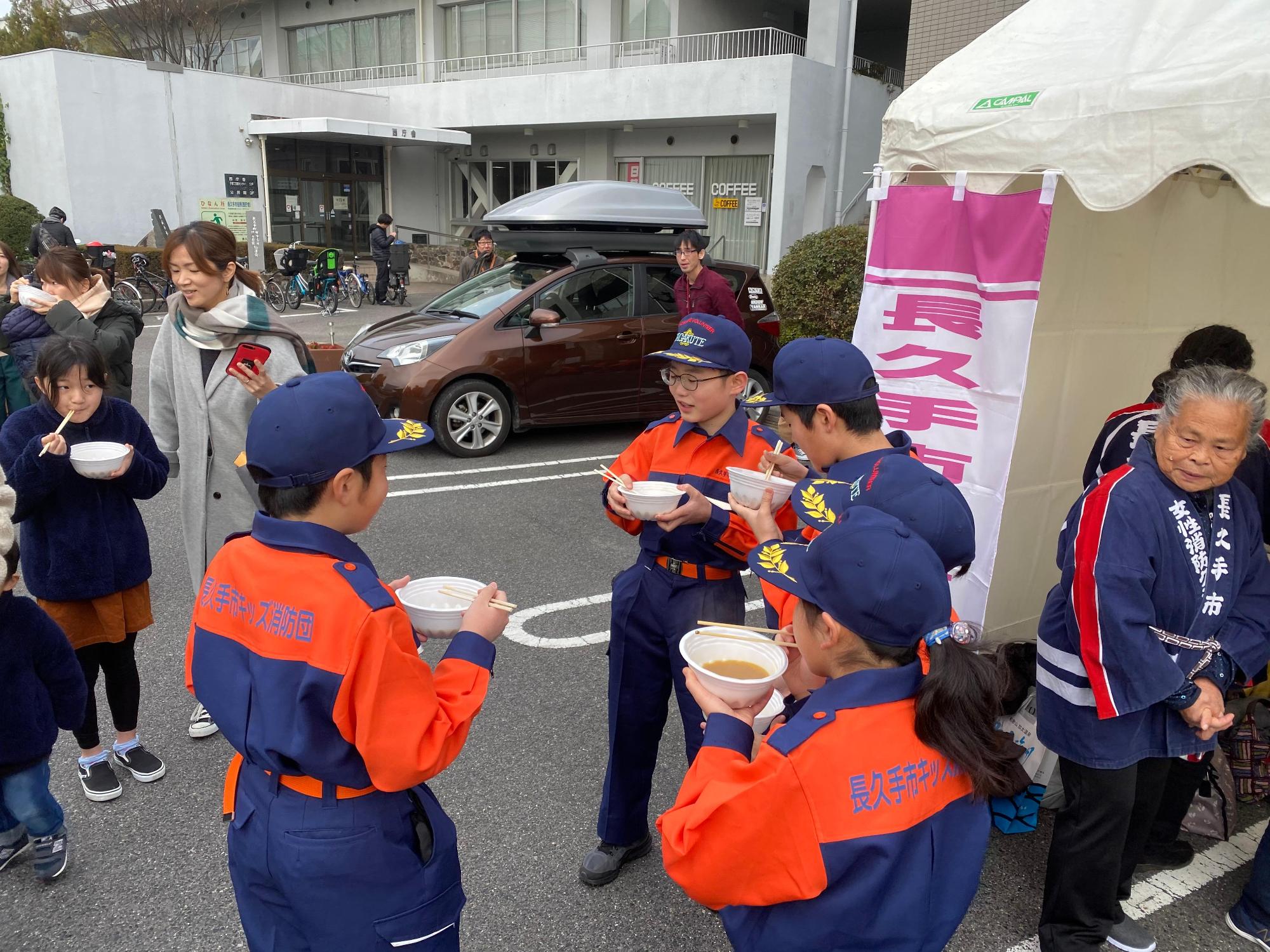 駐車場に設置されたテントの前で豚汁を試食する長久手市キッズ消防団員や来場者の写真