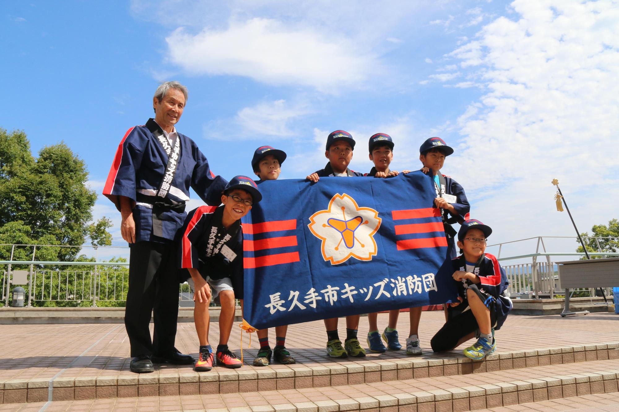 帽子を被り法被を着た男の子たちが消防団団旗を広げ記念写真を撮っている写真