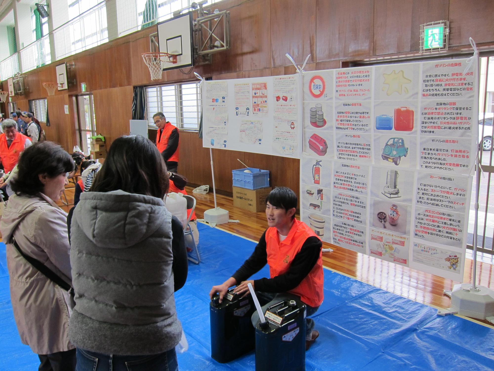 体育館の長久手市危険物安全協会のブースに携行缶の取扱についての資料が掲示されている前で、オレンジ色のベストを着たスタッフが携行缶を前に、参加者に話をしている写真