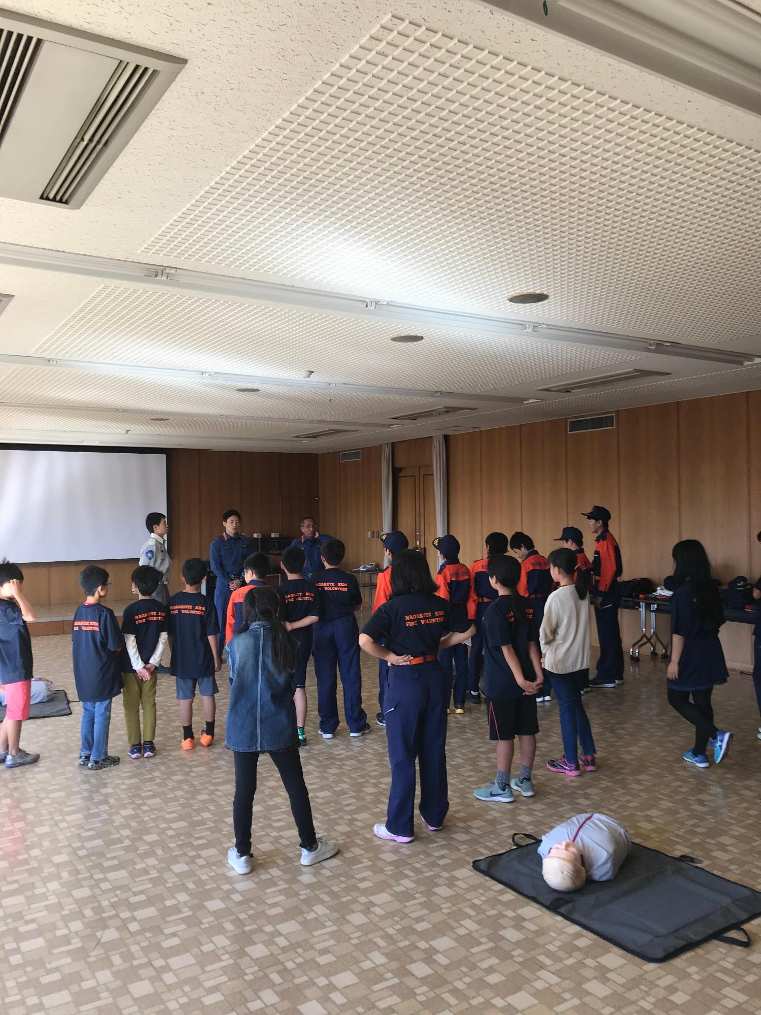 心肺蘇生用の練習用人形が横たわる室内で後ろに手を組んで整列している参加者達の写真