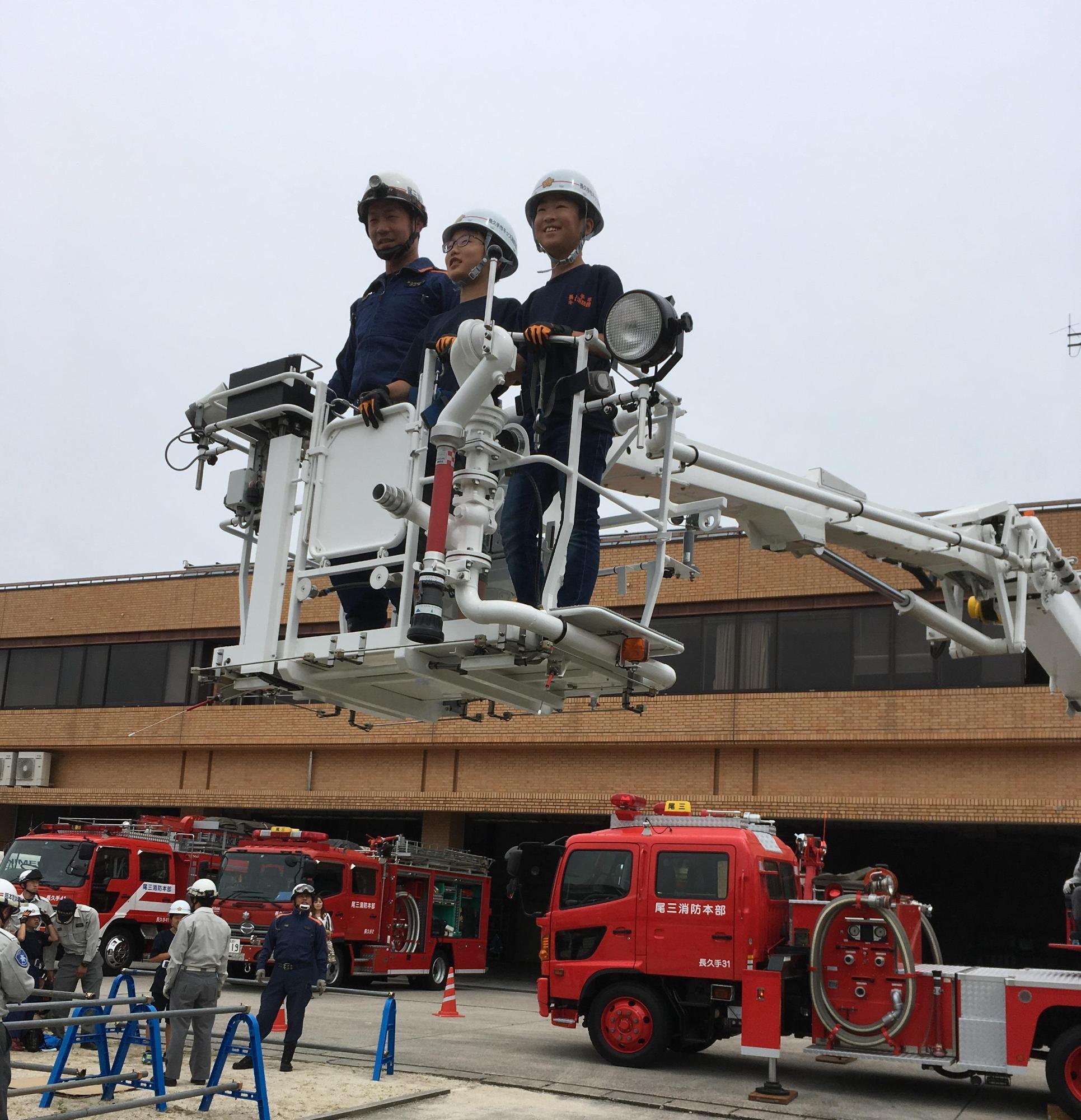 消防団員と一緒にはしご車に笑顔で乗っている長久手市キッズ消防団男子2名の写真
