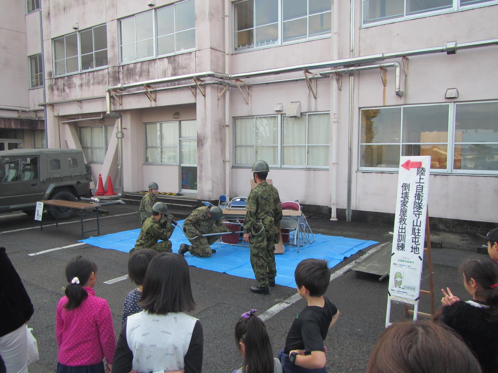 校舎の前で陸上自衛隊が人命救助訓練でテーブルの下から救出作業をしている様子を参加者たちが見ている写真