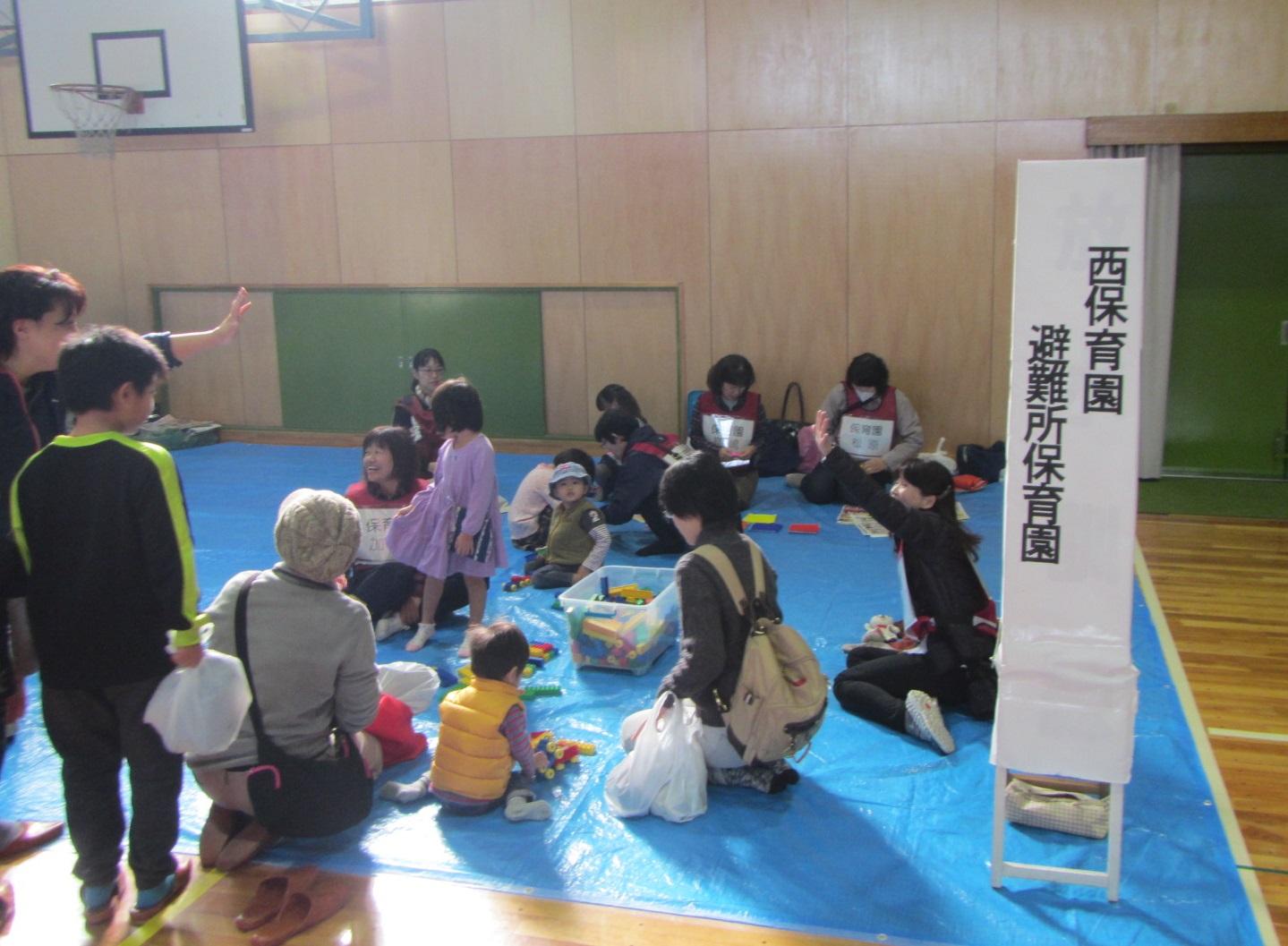 体育館の保育園のブースで保育士たちが子供たちとブロックなどで遊んたり、面倒を見ている写真