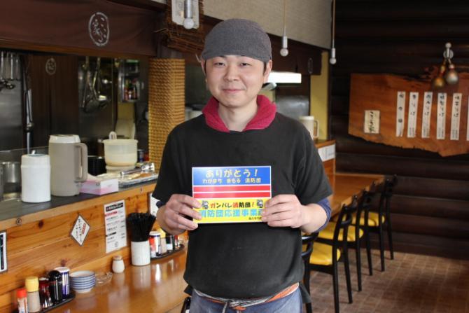 中華そば こころ家のご主人がカウンター席の前で消防団応援事業所表示証を持って写っている写真