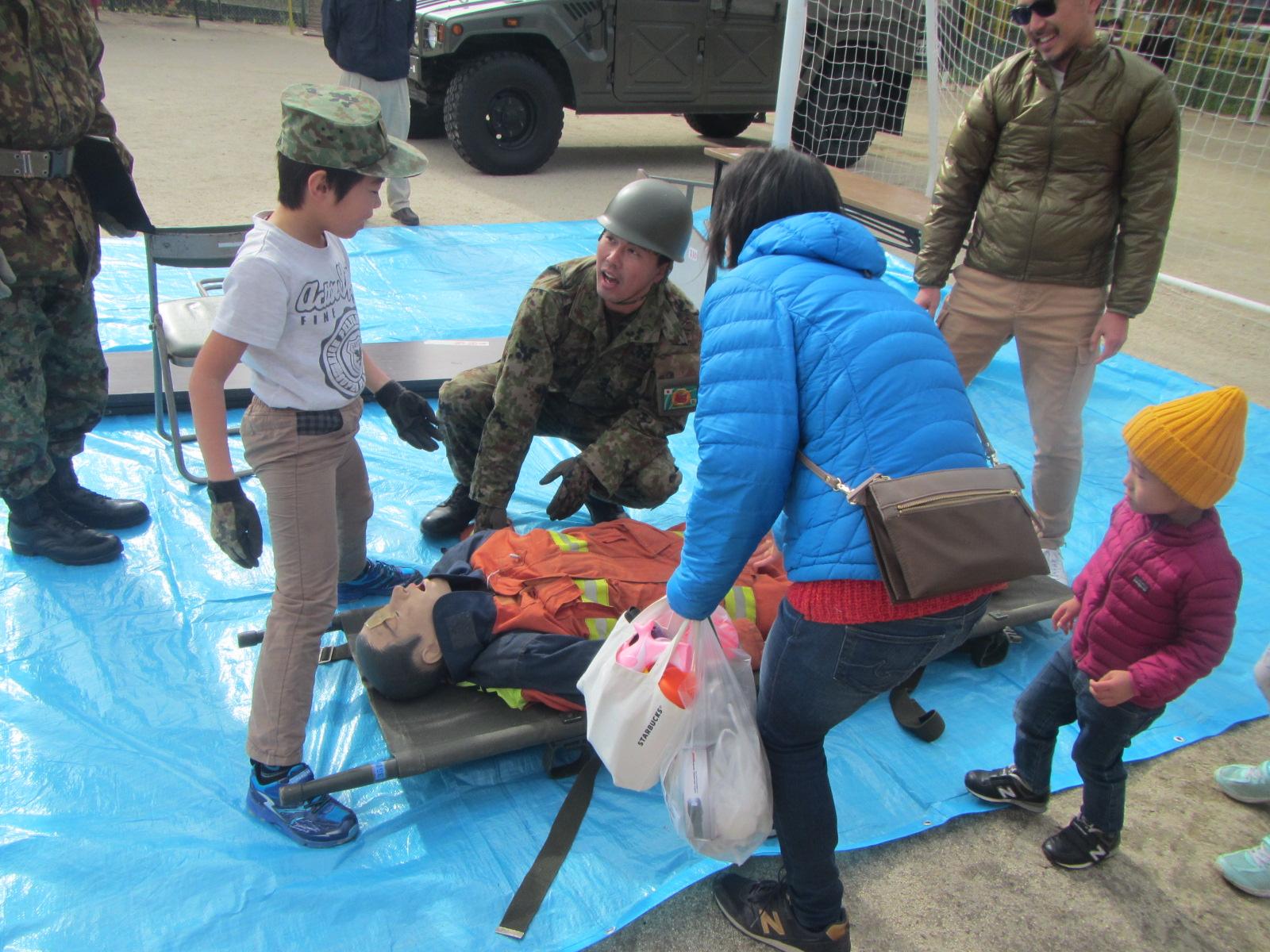 陸上自衛隊隊員の指導の下、担架に乗った人形を使って人命救助訓練をする参加者親子たちの写真