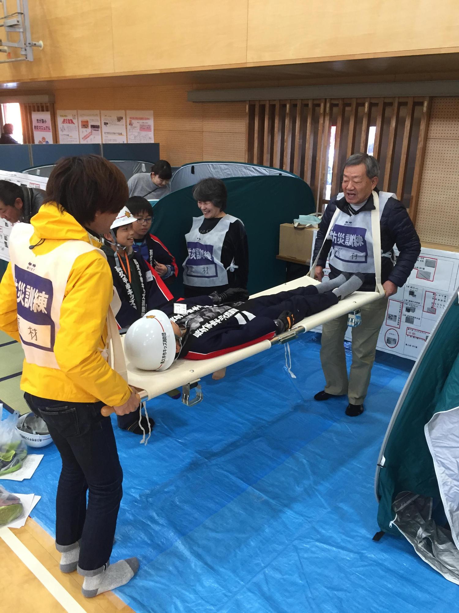 体育館でヘルメットに法被姿のキッズ団員が乗っている担架を訓練者が持ちあげている写真