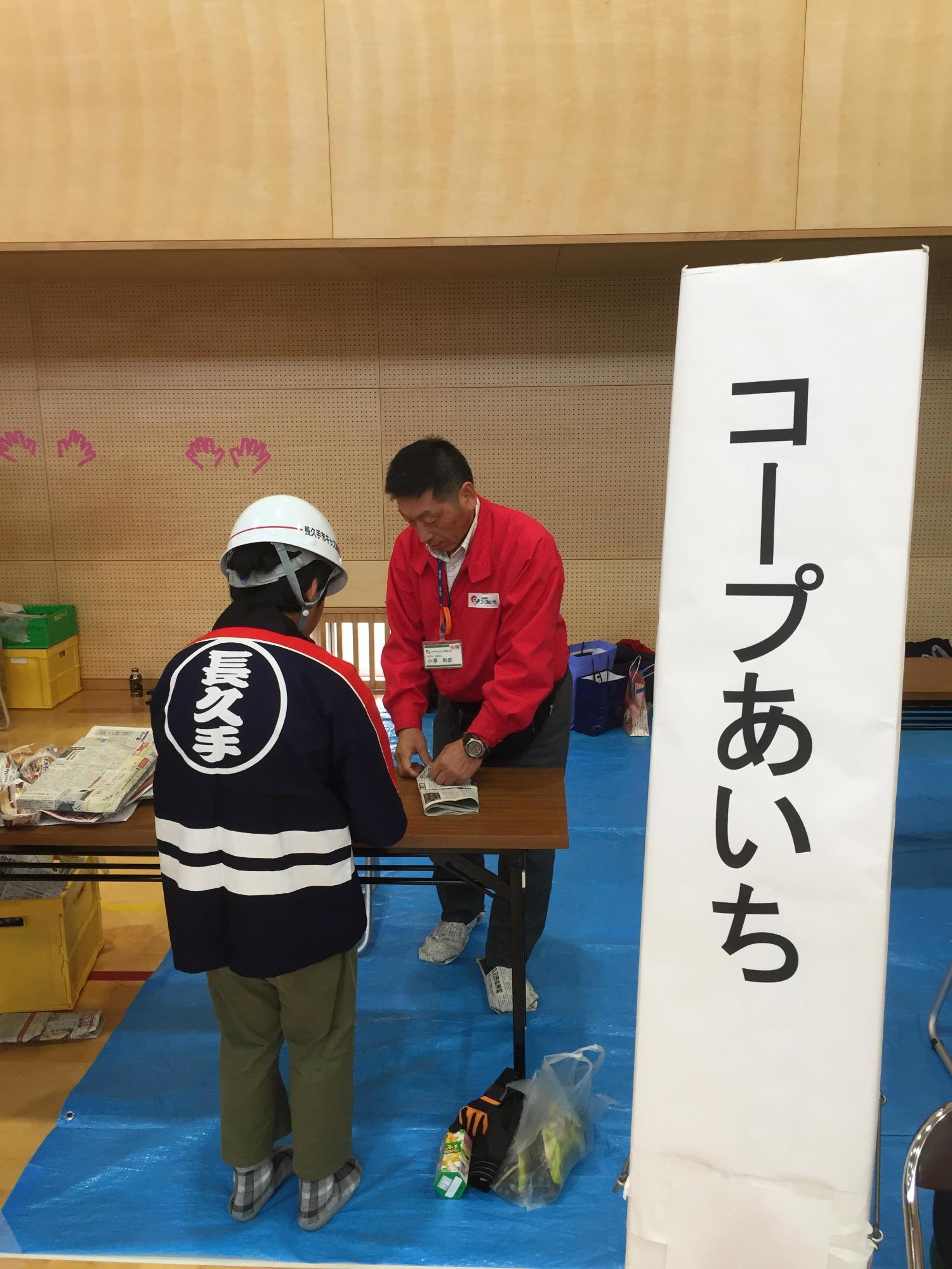 コープあいちのブースで赤いジャンバーを着たスタッフより新聞紙を使ったスリッパの作り方を教わるヘルメットに法被姿のキッズ団員の写真