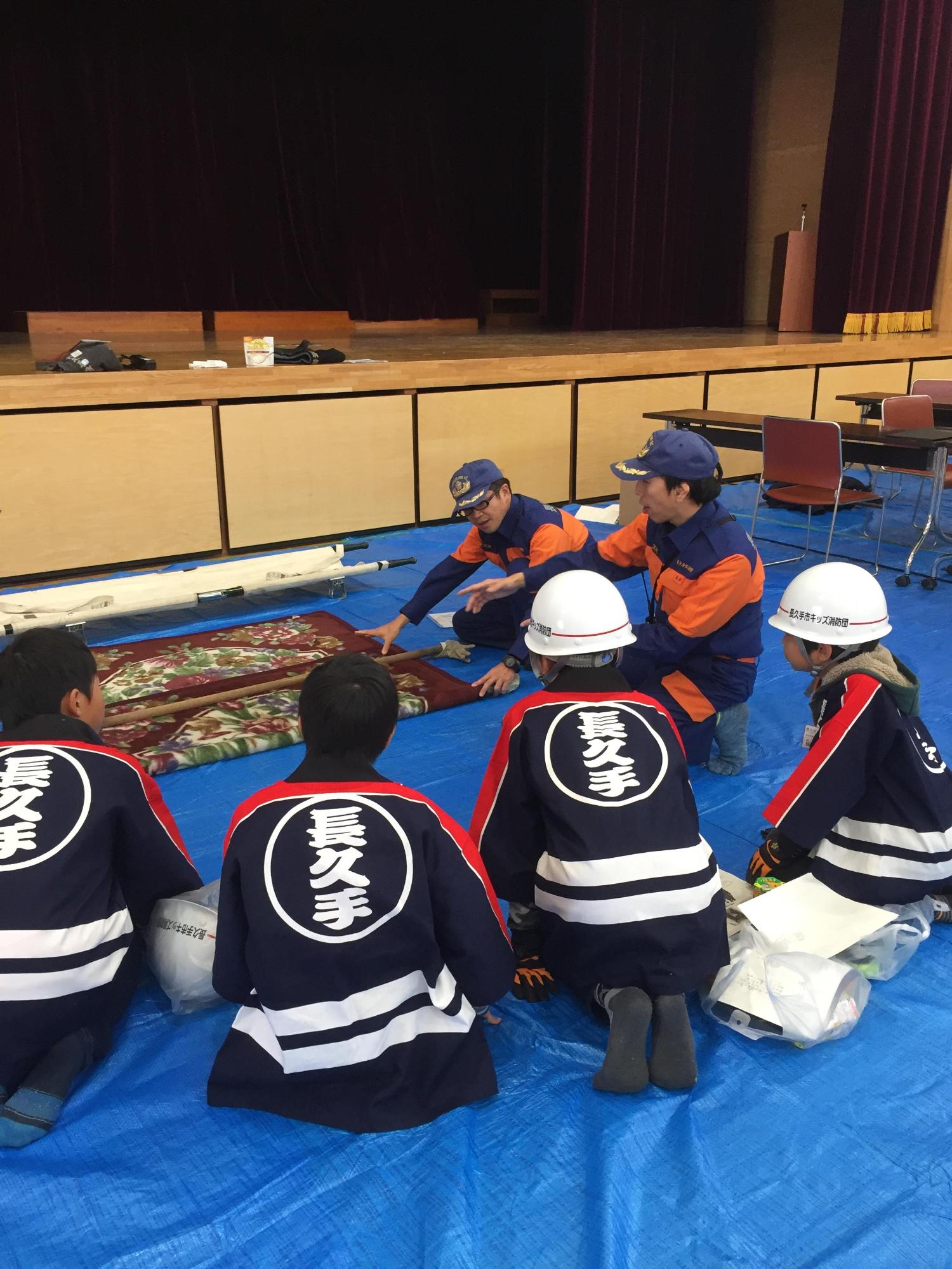 体育館で消防団員より担架の搬送について説明を受ける法被姿のキッズ消防団員たちの写真