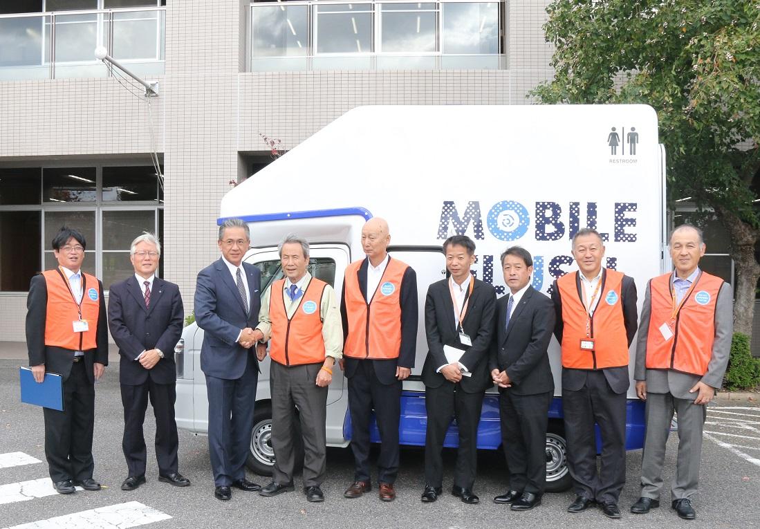 トイレカーの前で関係者9名の方が並んで記念撮影をしている写真