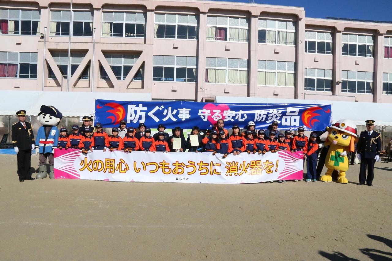 キッズ消防団員たちが火の用心の横断幕を持って学校の校舎の前でゆるキャラたちと記念撮影している写真