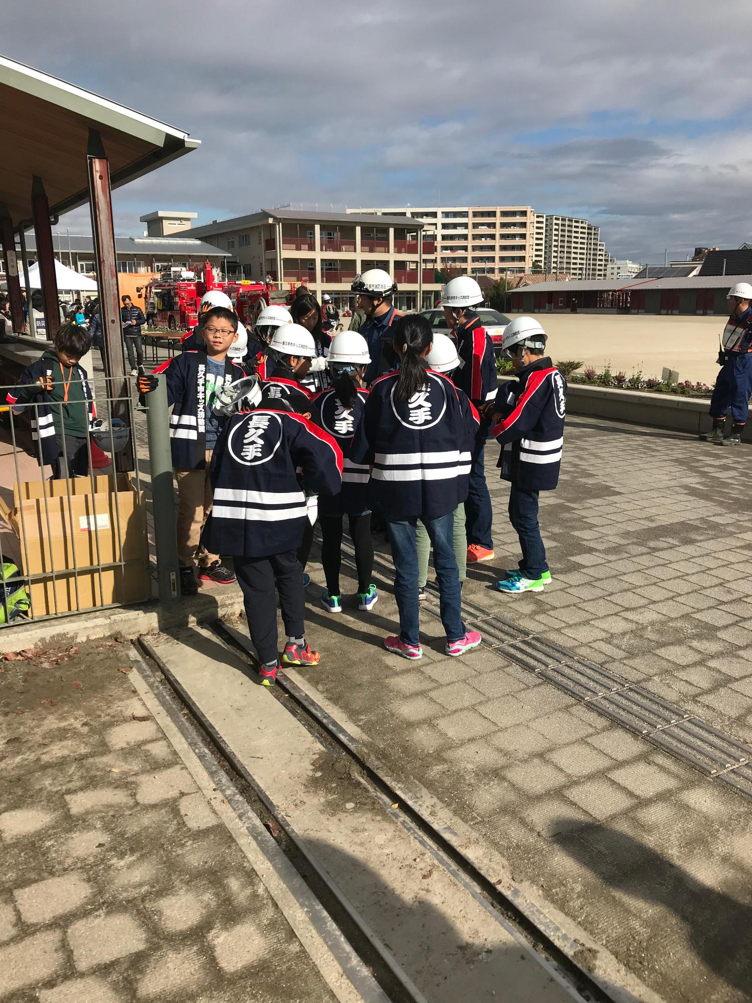 消防団員やヘルメットに法被姿のキッズ消防団員たちが集合している先にグラウンドや校舎が写っている写真