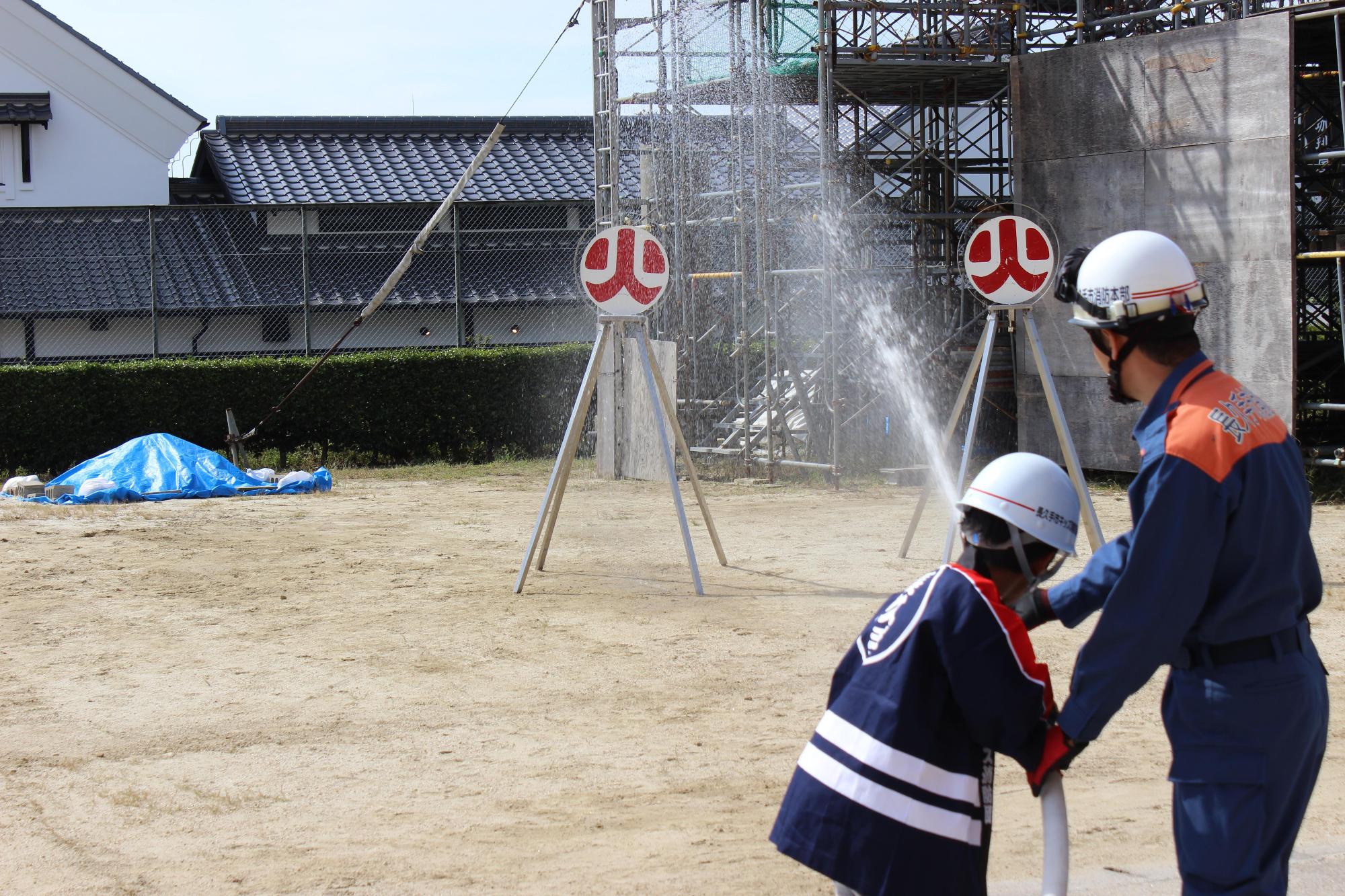 消防団員とキッズ消防団員が一緒にホースを持って放水している写真