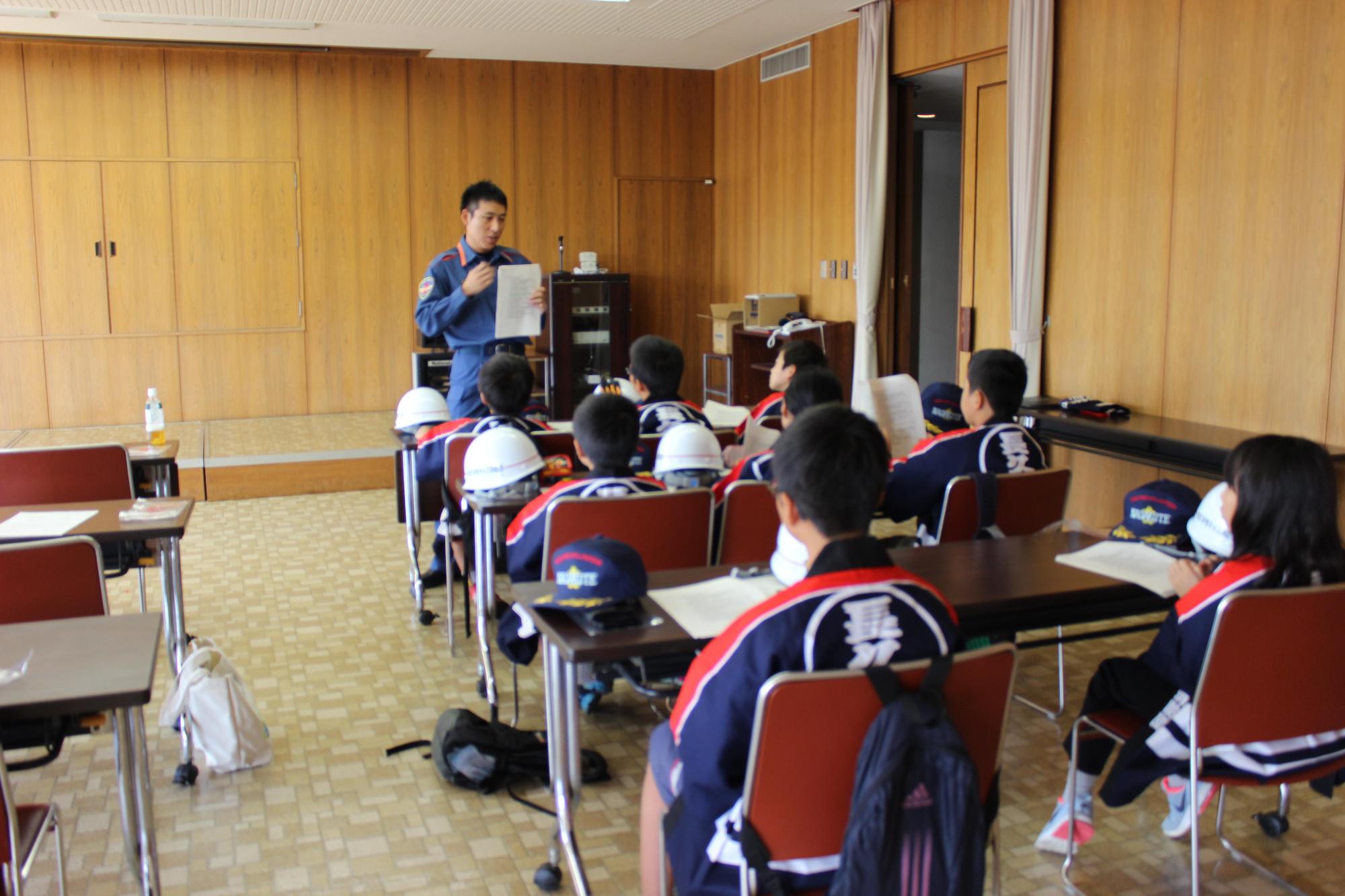 資料が並べられたテーブルに椅子に座り、消防署員より消防の歴史を学ぶキッズ消防団員たちの写真